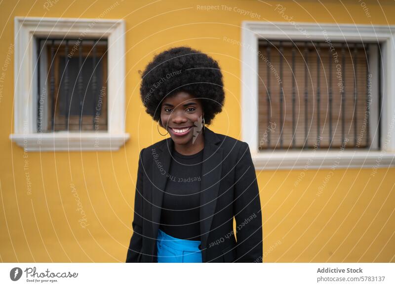 Smiling Afro Woman In Stylish Urban Attire woman afro hairstyle smile confident pose yellow wall urban lifestyle vibrant young stylish attire fashion portrait