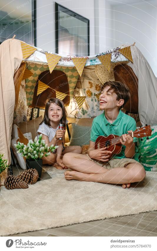 Happy children playing a ukulele and singing on handmade shelter tent in living room at home happy guitar laughing music teepee camping having fun diy indoor
