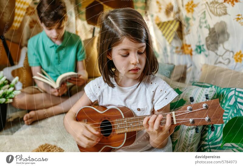 Portrait of concentrated little girl playing ukulele and boy reading book on handmade teepee at home portrait guitar children leisure shelter tent diy vacation