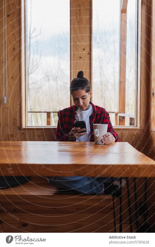 Casual woman using smartphone at wooden table in cabin mug window plaid shirt seated holding engrossed serene casual indoors technology mobile phone