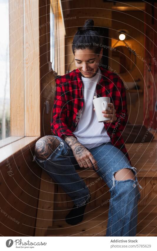 Casual moment of a tattooed woman enjoying coffee mug window smile wood interior casual cozy relaxed sitting holding enjoyment warmth beverage plaid shirt denim