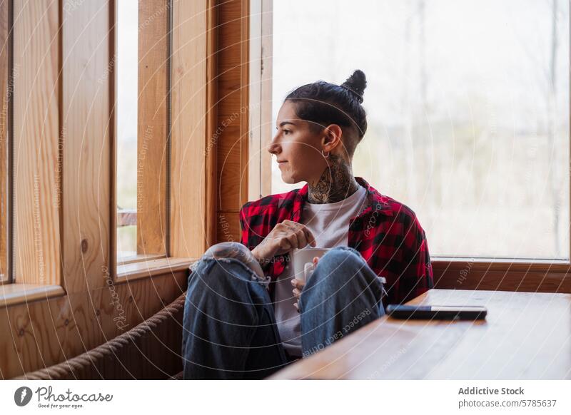 Contemplative tattooed individual with mug by window person contemplation serenity gaze indoor young thoughtful serene plaid shirt wood natural light calm