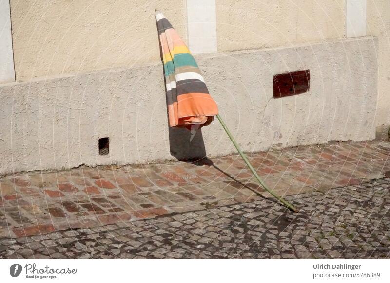 bent closed parasol with pale-colored stripes leaning against a house wall Sunshade quaint Humor Funny pale colours Street Shadow
