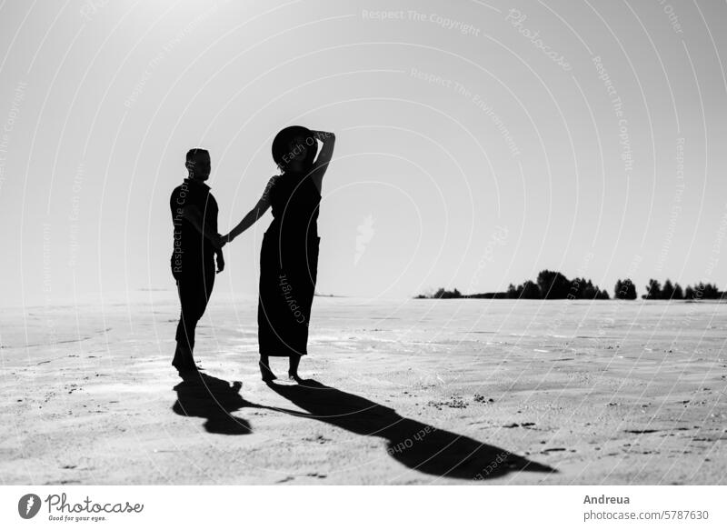 young couple a guy and a girl with joyful emotions in black clothes walk through the white desert barefoot blue day dress footprints freedom fun hat in the sand