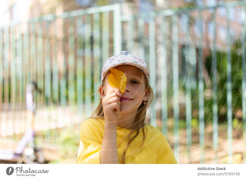 Girl with a yellow leaf covering one eye girl play smile cap park bright blurred background playful childhood outdoor innocence nature fall autumn foliage joy