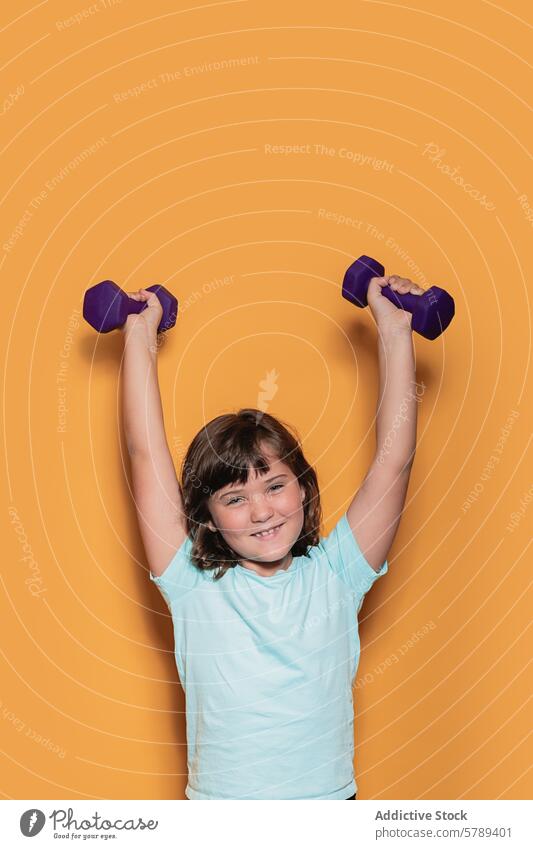 Cheerful child lifting weights on orange background fitness health sport exercise girl dumbbell studio smile joy happiness playful strength recreation sporty