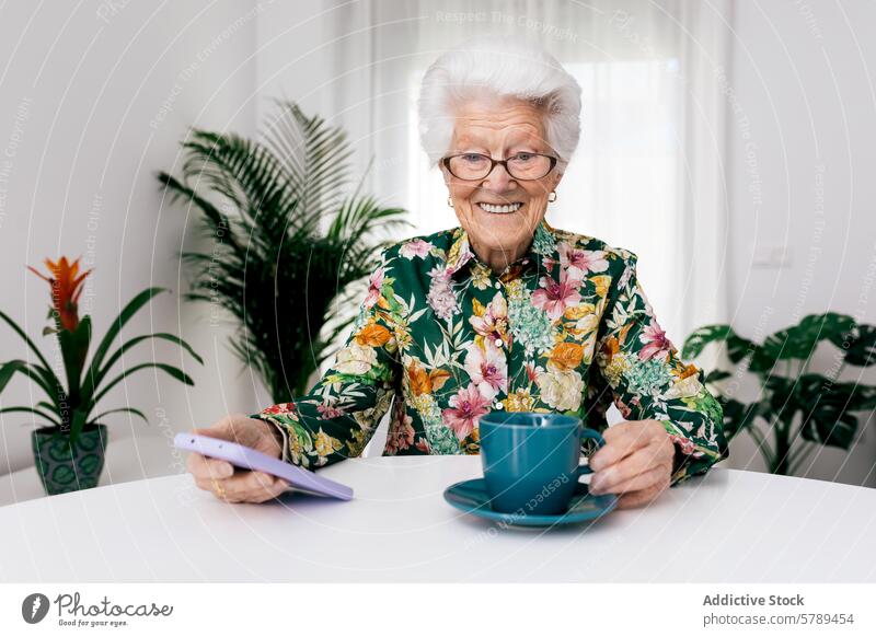 Elderly woman smiling at home with coffee and smartphone elderly senior glasses floral shirt cup drink holding table white houseplant cheerful enjoying indoors