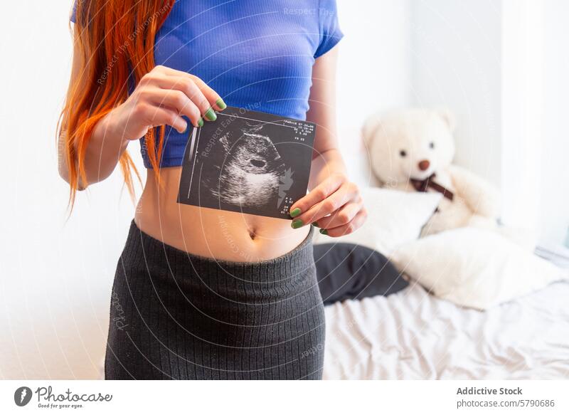 Expectant mother holding ultrasound image with teddy bear in background pregnancy maternity expectant woman young baby scan room cozy bed hair red showcase