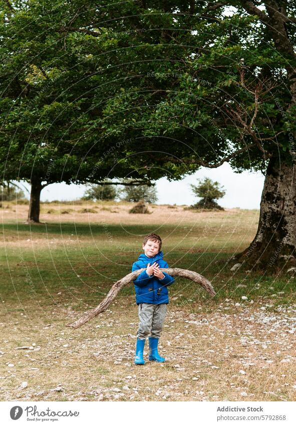 Young boy holds large stick in nature setting child outdoor tree field grass sky blue jacket boots playful adventure exploration leisure family kid happy youth