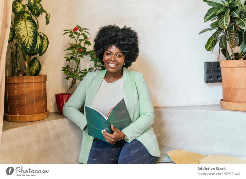 Smiling professional woman in a green blazer with a notebook smiling seated indoor plants coworking space style confidence positivity african descent cheerful