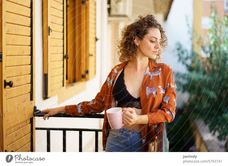 Young woman standing on balcony with cup of coffee casual terrace drink beverage curly hair calm peaceful lady dreamy alone hot drink leisure apartment building