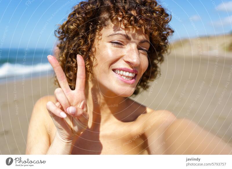 Happy woman showing V sign on beach selfie vacation smile summer v sign two fingers gesture cheerful happy young curly hair moment positive ocean chill sea