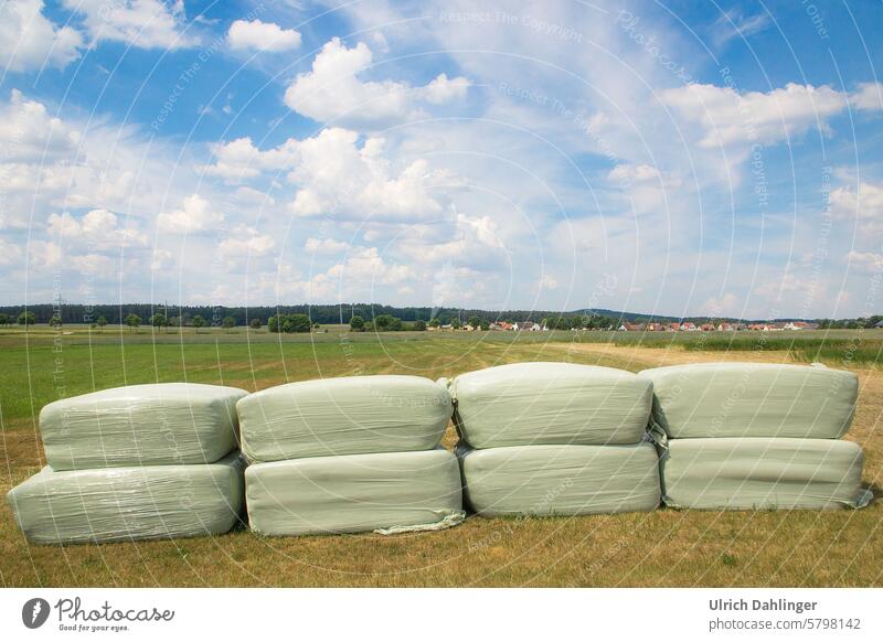 green meadow/field.in the background on the horizon forest and a village.half of the picture blue summer sky with white fair weather clouds.in the foreground eight hay bales in plastic packed to rectangular squares in pale green wrapping.