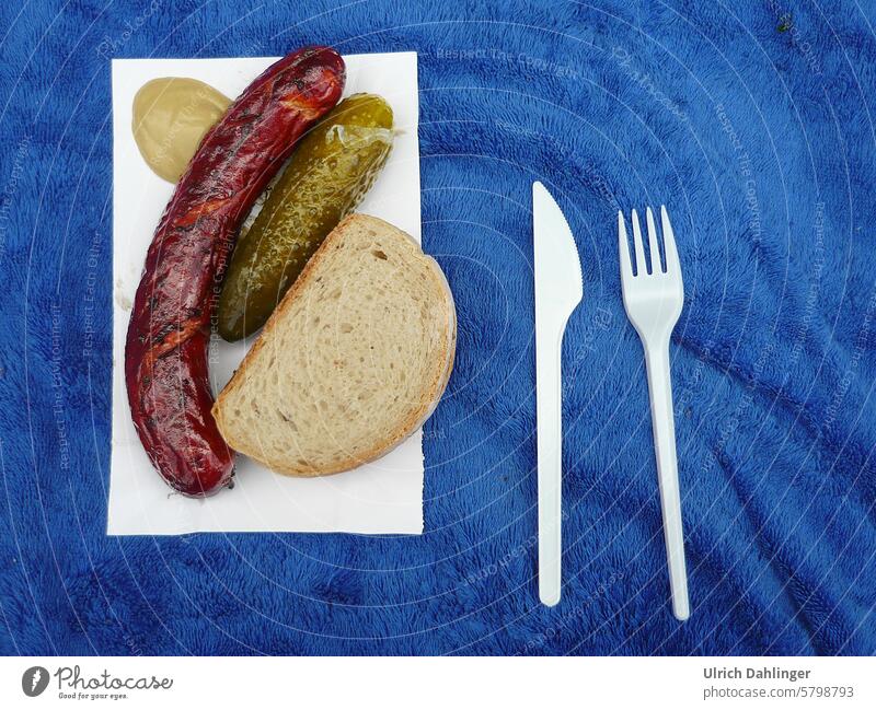 Bratwurst with bread, gherkin, mustard on white cardboard, plastic knife and fork next to it, all together on a blue terrycloth towel Snack bar