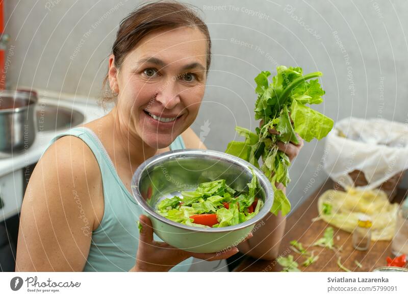 A woman shows a bowl of vegetables and herbs with lettuce leaves against the background of the kitchen. women female person salad food lifestyle vegetarian