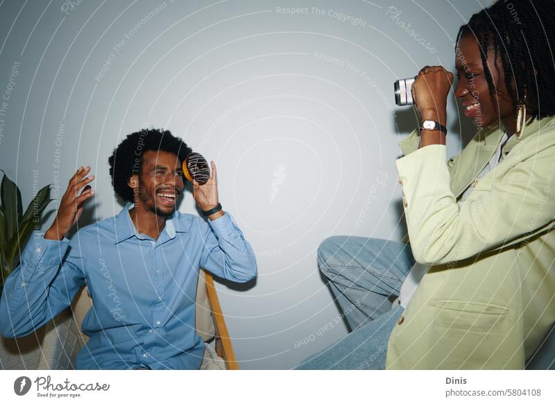 Smiling creative Black woman photographing coworker in office posing fun Party doughnut playful happy celebrate Friday businesswoman colleague black man