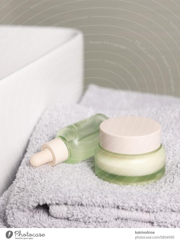 Light green dropper bottle and cream jar on light grey bath towel near basin closeup Green mockup Dropper Cream Bath Basin Cosmetic Beauty Skincare routines