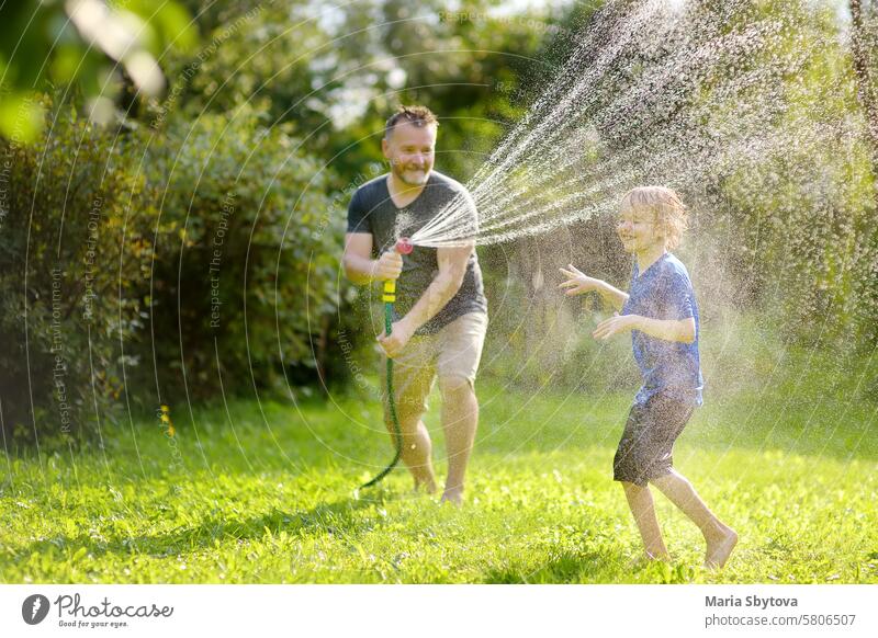 Funny little boy with his father playing with garden hose in sunny backyard. Preschooler child having fun with spray of water. Summer outdoors activity for kids.