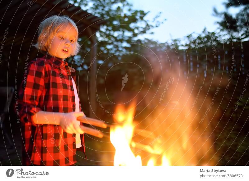 Cute little boy is burning a bonfire on a summer evening in the backyard. Child puts firewood in a fire bowl by tongs. Summer holidays for kids in village.