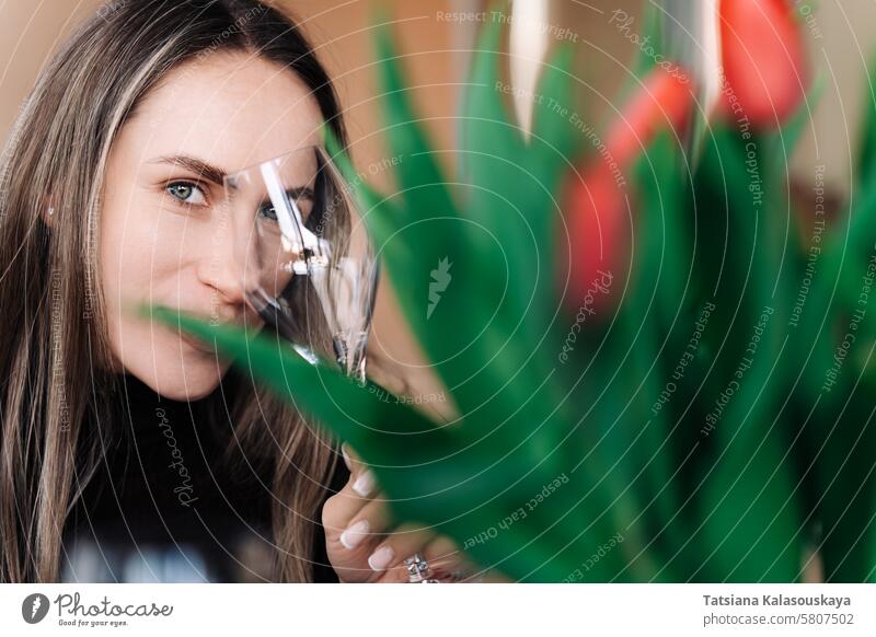 Young woman looks mysteriously at camera against the background of a blurred bouquet of flowers, covering one eye with a wine glass women springtime humor