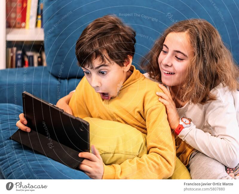 Kids Engaged in Digital Tablet on Cozy Blue Sofa children two surprised kids tablet sofa cozy digital technology home indoor learning entertainment play