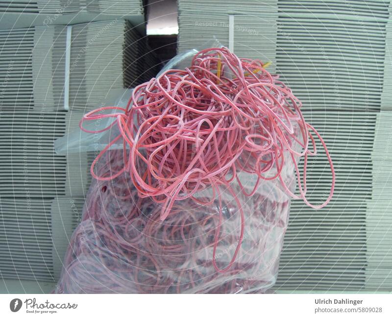 Red rubber bands spilling out of a plastic bag, with stacks of gray cardboard boxes in the background rubber rings Packaging Cardboard Packaging material