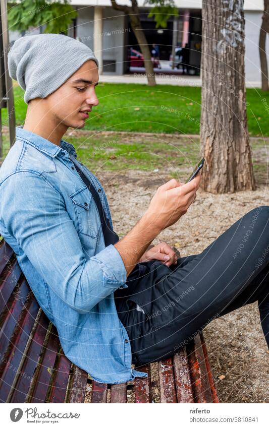 Man in denim and beanie using smartphone on bench. young man jacket park focused trees grass casual outdoor sitting technology mobile urban lifestyle