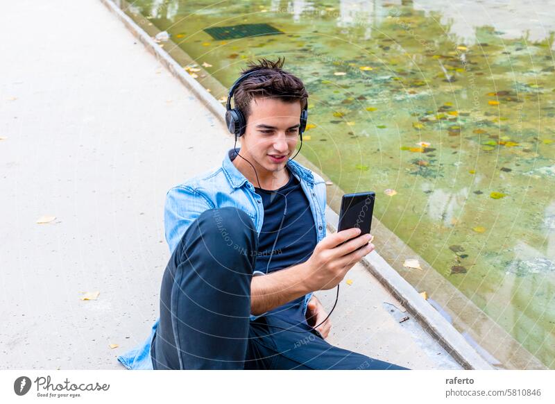 Man listening to music by a pond. man headphones smartphone denim shirt black pants young adult focused relaxed urban outdoor sitting casual technology mobile