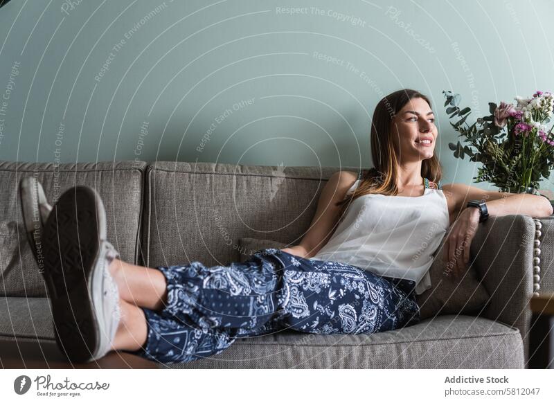 Dreamy woman resting on sofa in house dreamy smile legs crossed chill charming admire couch living room content enjoy contemplate gadget device lifestyle