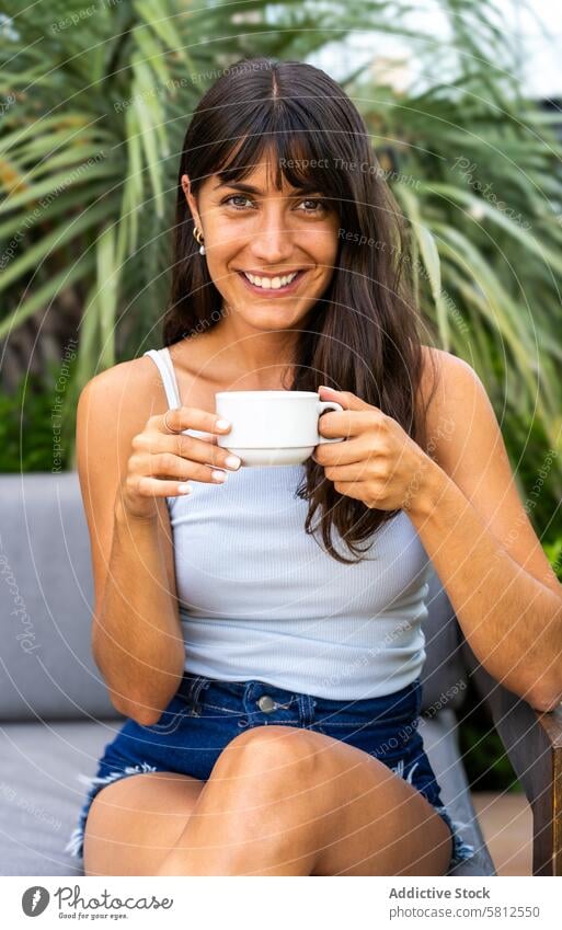 A woman is lookintg at camera while sitting on a couch and holding a white cup. She is smiling and she is enjoying her coffee Modern adult attractive background