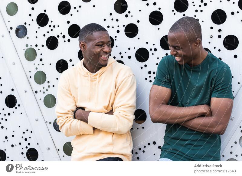 Cheerful black friends with folded arms near ornamental wall arms crossed laugh spend time weekend toothy smile masculine town casual spare time arms folded