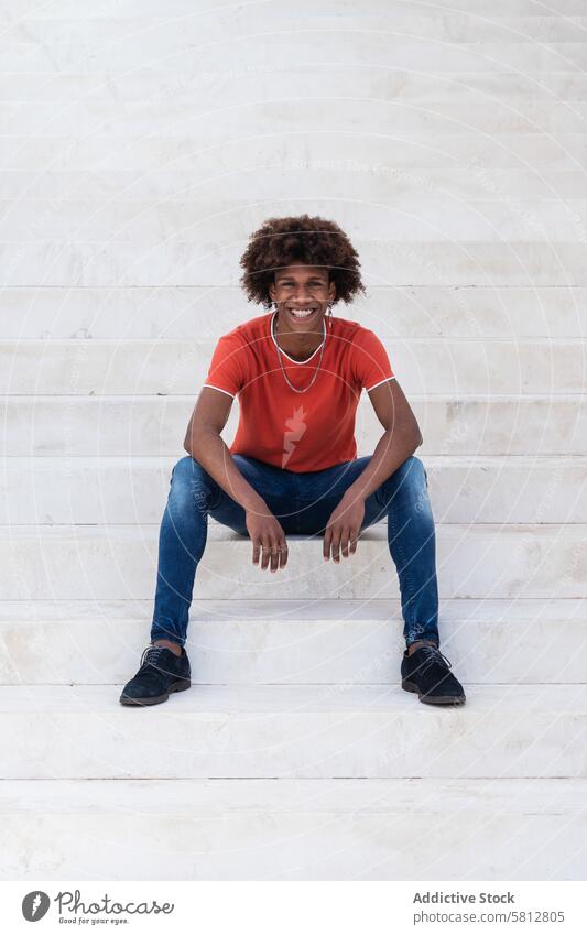 Happy young African American man sitting on staircase and smiling rest smile delight street happy style cool appearance chill content male ethnic