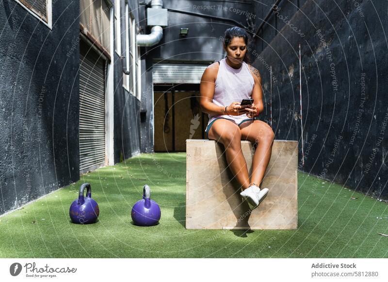 Ethnic woman browsing smartphone on jump box using fitness training kettlebell break workout female serious gym wooden sportswear gadget focus strong wellness