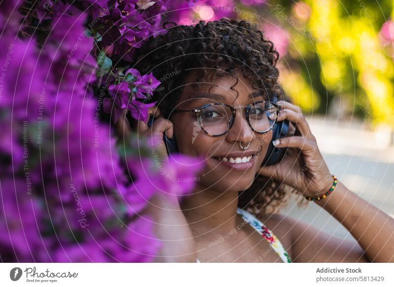 Black woman listening to music near flowers bush garden headphones smile style young female glasses piercing ethnic black african american song joy trendy