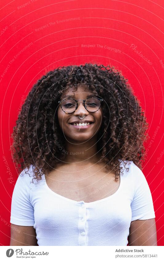 Happy African American female against red wall woman style smile appearance color bright curly hair glasses piercing ethnic black african american cheerful