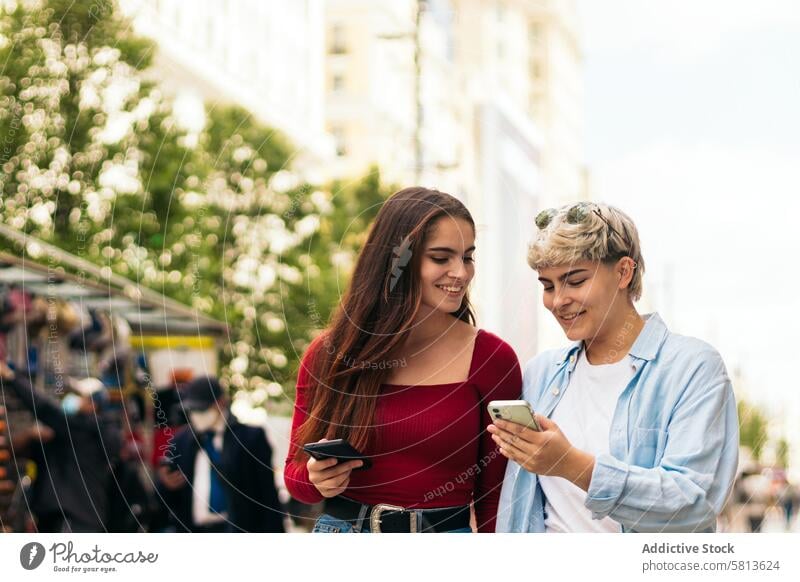two teenager girls walking and watching mobile phone looking smartphone holding laughing smiling internet app outdoor happy young woman cellphone people