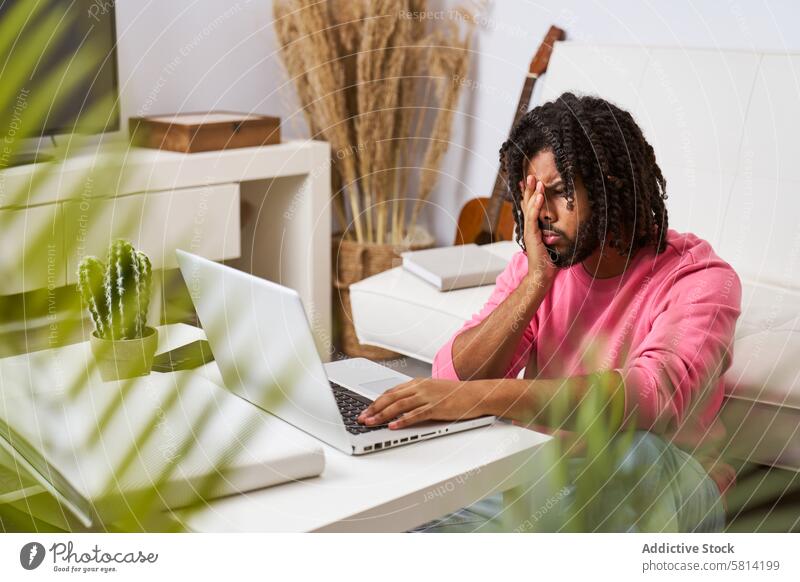 worried and depressed man working from home covering his face with his hands in front of the laptop young receiving bad news hairstyle braided computer internet