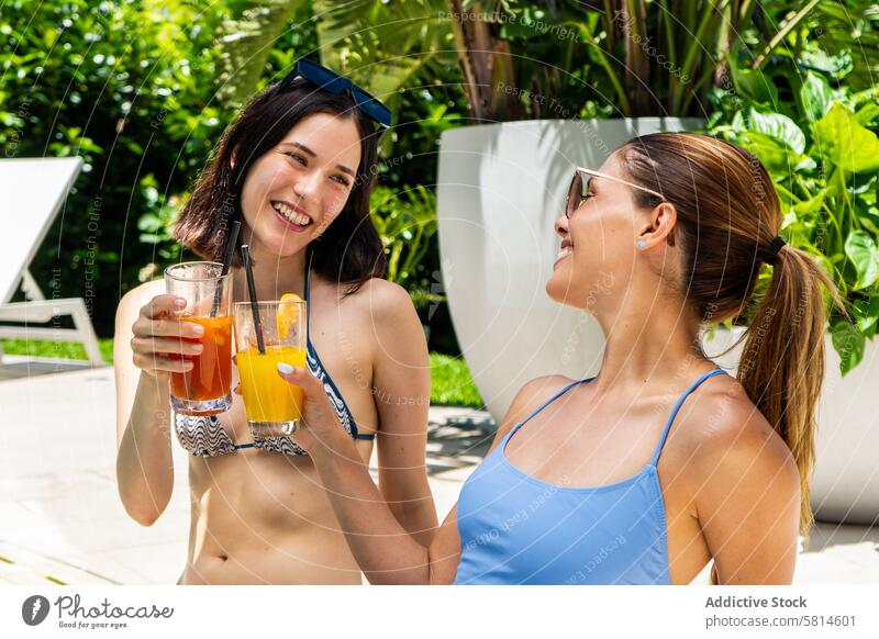Two friends sitting by a swimming pool, each holding a fruit smoothie. attractive beautiful best beverage bikini bonding carefree chair courtyard deck chair