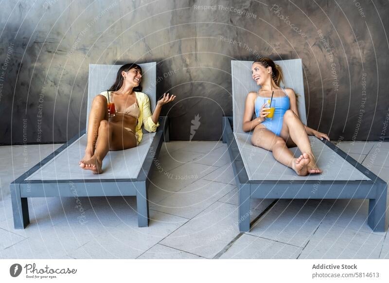 Front view of two women friends resting on lounge chairs while talking. adult attractive beautiful beauty best beverage bikini bonding carefree caucasian