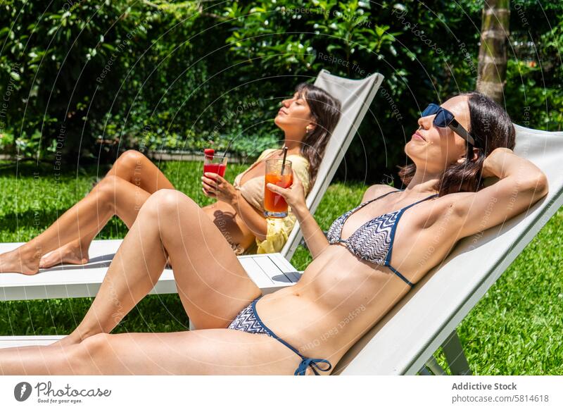 Two friends sunbathing on deck chairs holding fruit smoothies attractive beautiful best beverage bikini bonding carefree courtyard drinking enjoying enjoyment