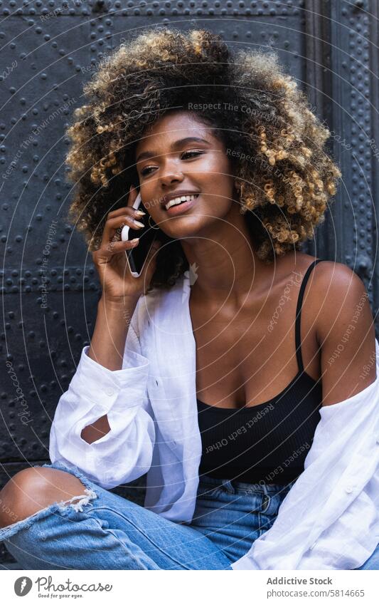 Delighted black woman speaking on smartphone while sitting near building positive phone call using communicate glad conversation pleasure female happy gadget