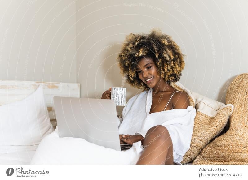 Black woman drinking coffee while sitting on bed with laptop using morning comfort bedroom happy chill female attractive apartment curly hair online gadget
