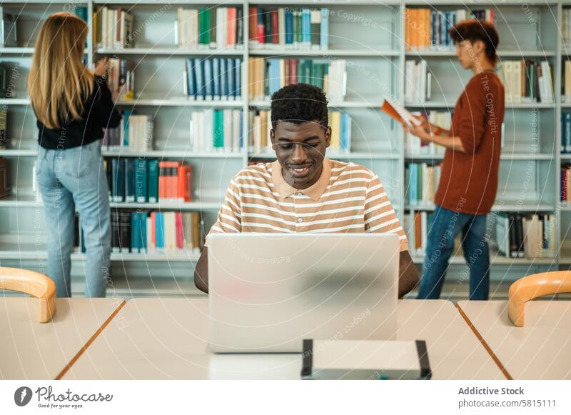 Black student browsing laptop in library using homework assignment smile study classmate bookcase pick teen adolescent teenager young education bookshelf