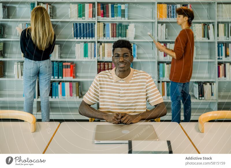 Happy young black man sitting with closed laptop student library smile positive bookshelf bookcase education study university male device gadget netbook