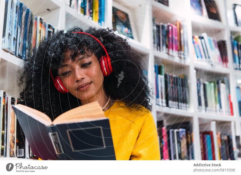 Content ethnic female student reading textbook in library woman smile education happy bookworm bookcase positive trendy young curly hair casual knowledge style