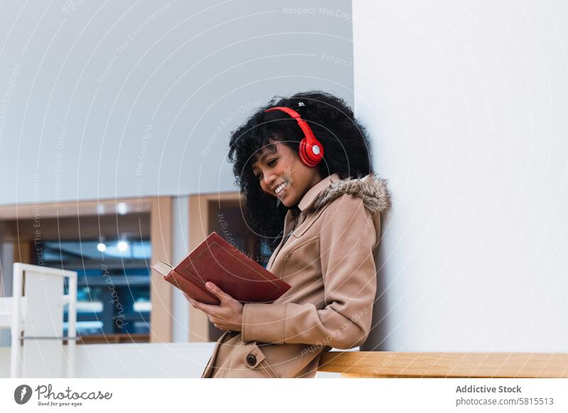 Content ethnic female student reading textbook in library woman melomaniac shelf bookcase headphones positive choose bookshelf smile music listen education