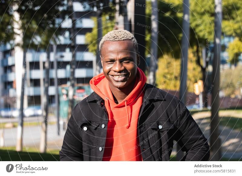 Confident young black man smiling in city park in sunlight smile rest positive style tree cheerful happy joy glad male african american ethnic human face