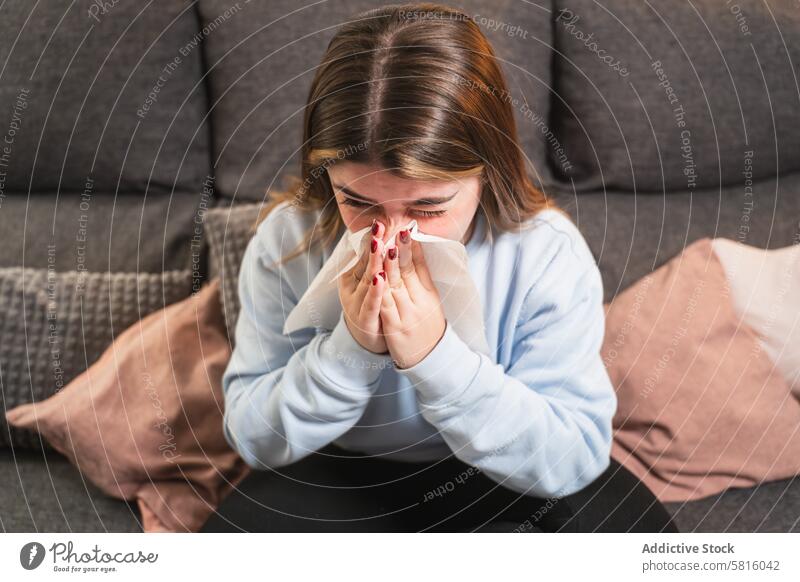Young woman with a cold sitting on a couch at home tissue sickness blowing nose allergy indoor female young illness health care flu symptom living room comfort
