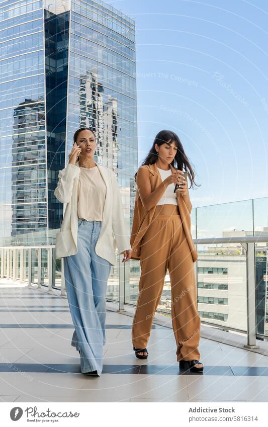 Two businesswomen are walking on a glass walkway. One of them is talking on the phone, and the other is sending a message. Beautiful People Modern attractive
