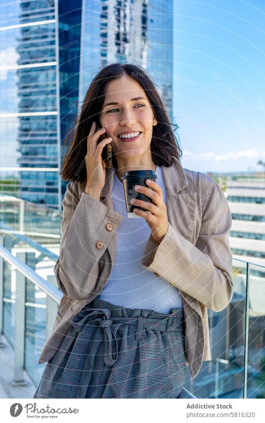 A woman is standing on a balcony and talking on her cell phone on her office break.  She has a paper cup of coffee in her hand. Modern attractive background